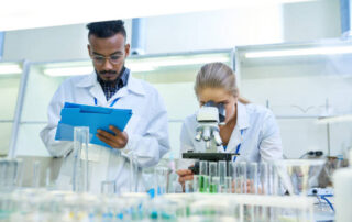 Portrait of young Middle-eastern scientist taking notes on clipboard while working on medical research in laboratory while his female colleague looking in microscope