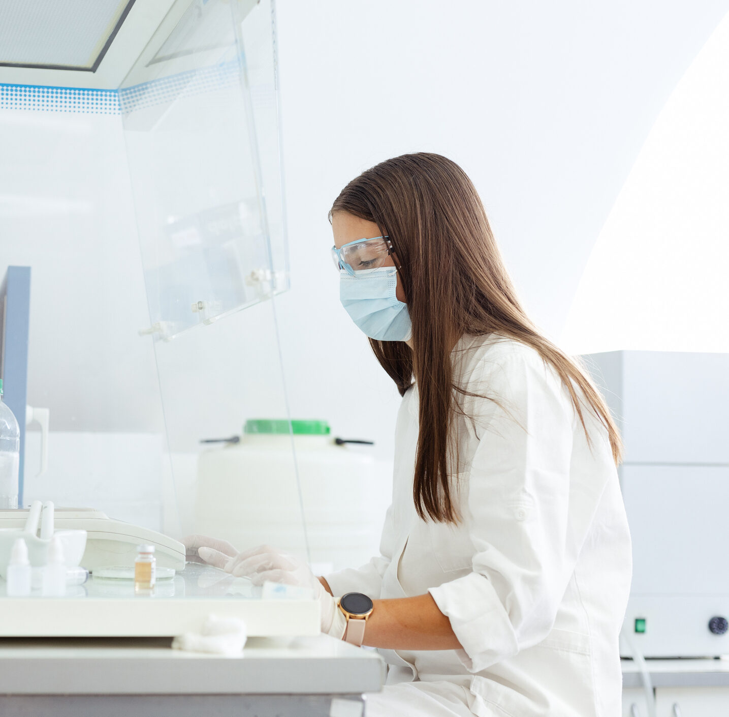 Scientist conducting lab research with modern equipment. A scientist in a white lab coat working at a laboratory bench, surrounded by scientific instruments, glassware, and a sterile laminar flow hood, indicating a research environment.